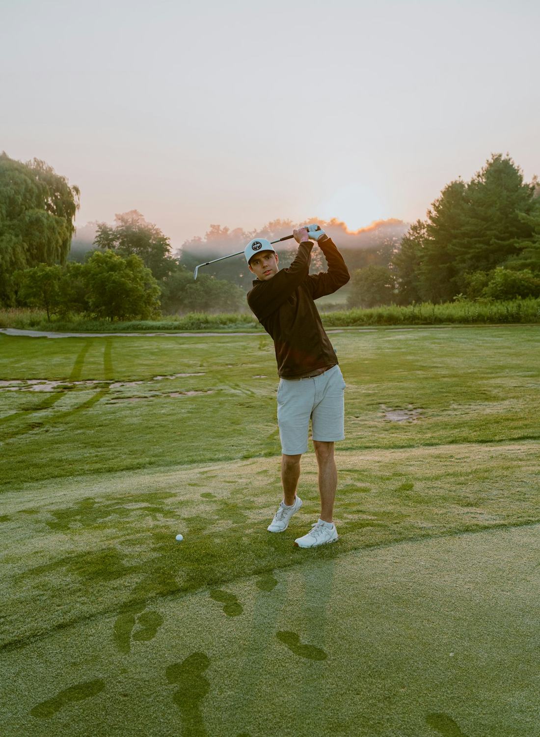 A golfer mid-swing on a green fairway in low evening light